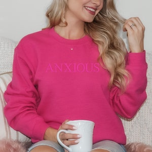 May include: A pink sweatshirt with the word "ANXIOUS" printed in pink on the front. The sweatshirt is being worn by a person holding a white mug.