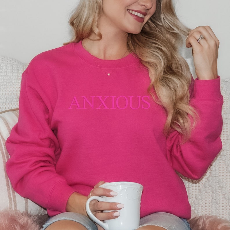 May include: A pink sweatshirt with the word "ANXIOUS" printed in pink on the front. The sweatshirt is being worn by a person holding a white mug.