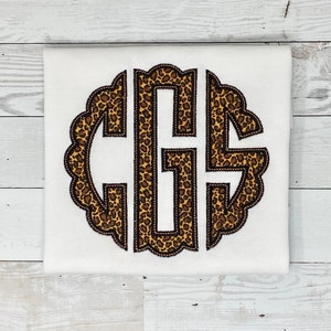 May include: A white shirt with a brown and black leopard print monogram in a scalloped circle.
