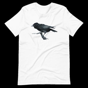 Puede incluir: Una camiseta blanca con una ilustraci&oacute;n de un cuervo negro impresa en la parte delantera. El cuervo est&aacute; posado en una rama y mira hacia la derecha. La ilustraci&oacute;n est&aacute; hecha en estilo acuarela.