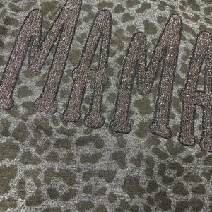 May include: Close-up of a dark grey fabric with a leopard print pattern. The word "MAMA" is embroidered in a glittery, dark grey thread across the fabric. The letters are in a cursive style.
