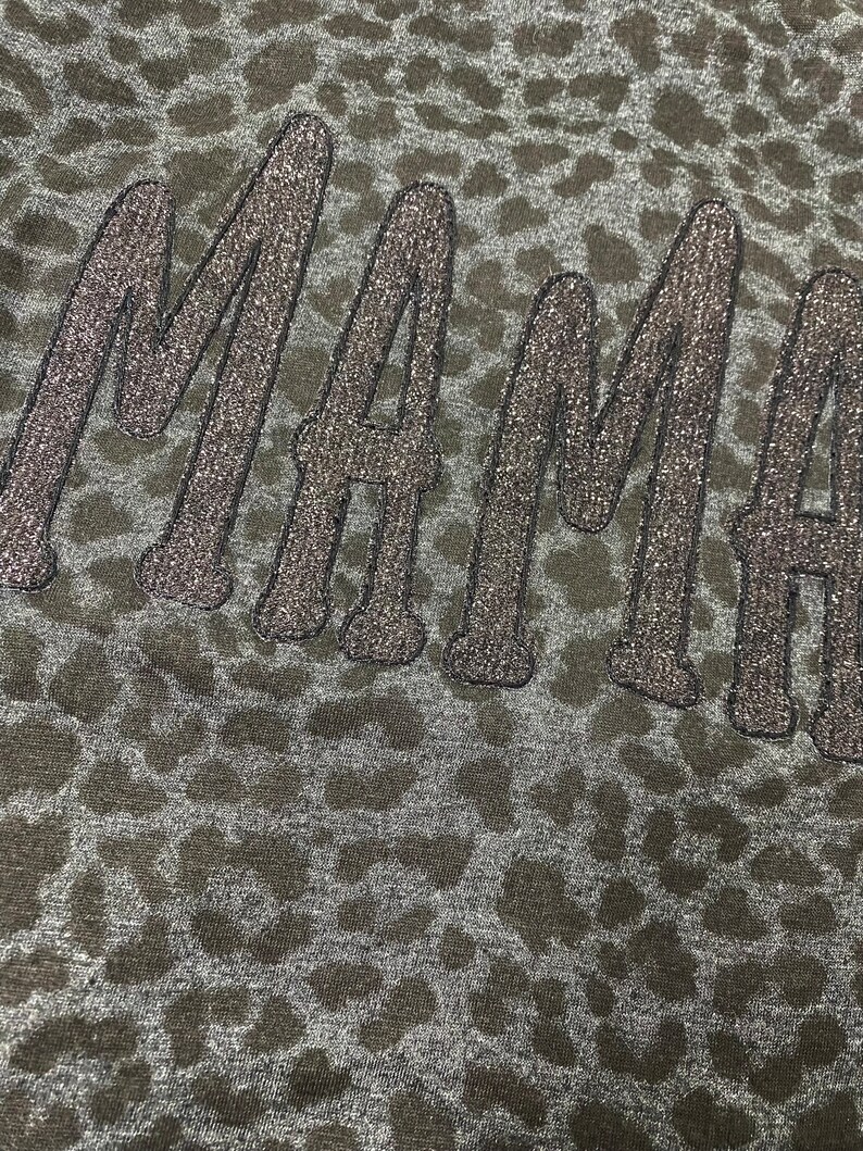 May include: Close-up of a dark grey fabric with a leopard print pattern. The word "MAMA" is embroidered in a glittery, dark grey thread across the fabric. The letters are in a cursive style.