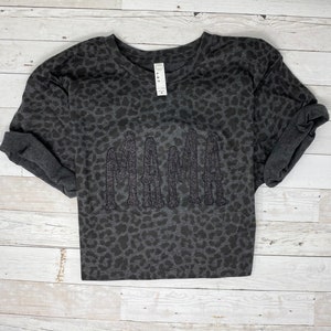 May include: A dark grey long-sleeved sweatshirt with a leopard print pattern. The word "MAMA" is embroidered across the front in a glittery, textured font. The sleeves are rolled up. The shirt is displayed on a light wood background.