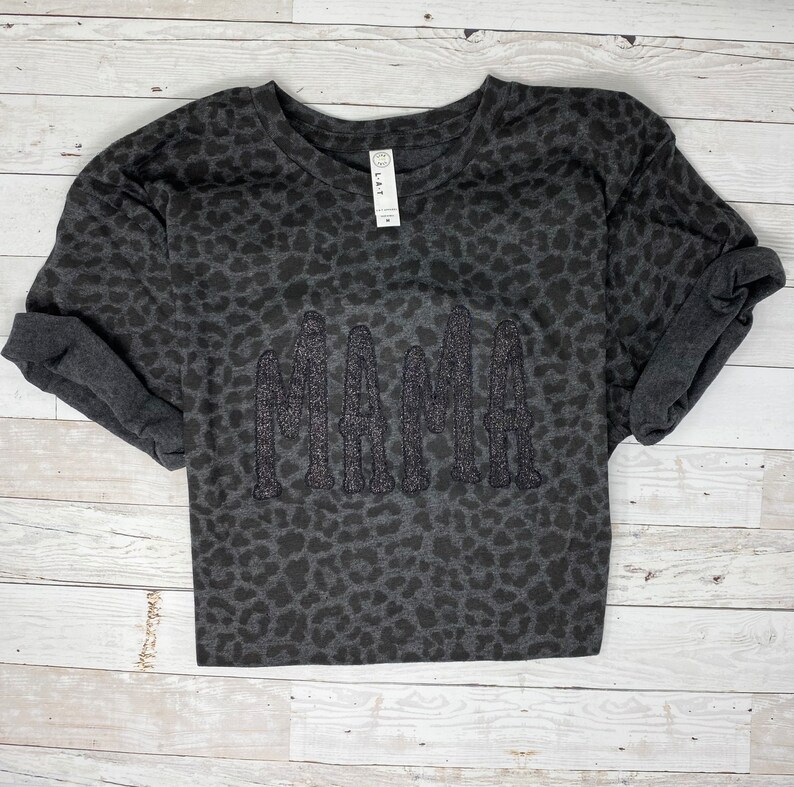 May include: A dark grey long-sleeved sweatshirt with a leopard print pattern. The word "MAMA" is embroidered across the front in a glittery, textured font. The sleeves are rolled up. The shirt is displayed on a light wood background.