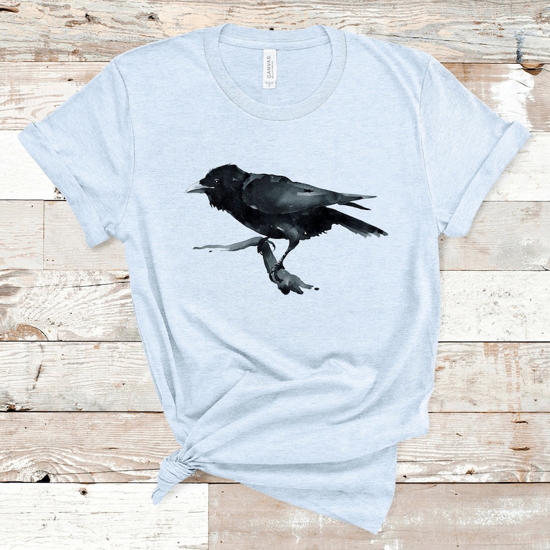 Puede incluir: Una camiseta azul claro con una ilustraci&oacute;n de un cuervo negro en acuarela. El cuervo est&aacute; posado en una rama y tiene un dise&ntilde;o detallado.