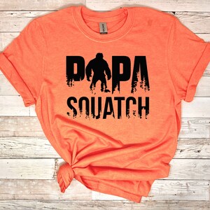 May include: Orange t-shirt with a black graphic of a Bigfoot with the text "DAPA SQUATCH" printed on it.