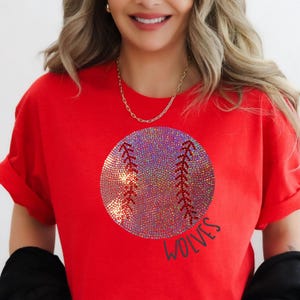 Pode incluir: Uma camiseta vermelha com um design de beisebol e a palavra "WOLVES" em preto. O gráfico de beisebol é iridescente. A camisa tem mangas enroladas e gola redonda.