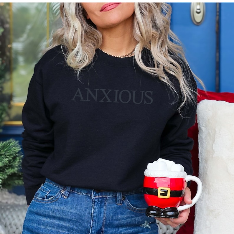 May include: A black sweatshirt with the word "ANXIOUS" printed on the front. The person is wearing blue jeans and holding a red Santa mug with marshmallows.