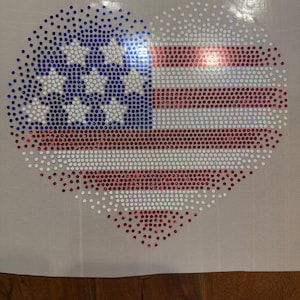 May include: Heart-shaped design featuring the American flag, crafted with red, white, and blue rhinestones. The design includes the stars and stripes of the flag, arranged in a heart shape. The rhinestones sparkle and catch the light.