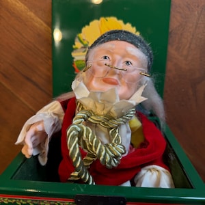 May include: A vintage doll of an elderly woman with glasses, wearing a red coat and a white ruffled collar, sits inside a green box. The box has a decorative clasp and a floral design on the lid. The doll has a gray hat and long white hair.