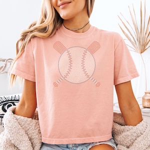 Peut inclure: T-shirt rose clair avec un motif de baseball. Le ballon est blanc avec des détails rouges et bleus, croisé par deux battes rouges. Le t-shirt a des manches courtes et un col rond.