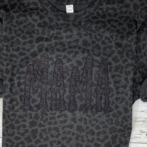 May include: Dark grey sweatshirt featuring a leopard print design. The word "MAMA" is embroidered in glittery, dark grey thread across the front. The sweatshirt has a crew neck and long sleeves.