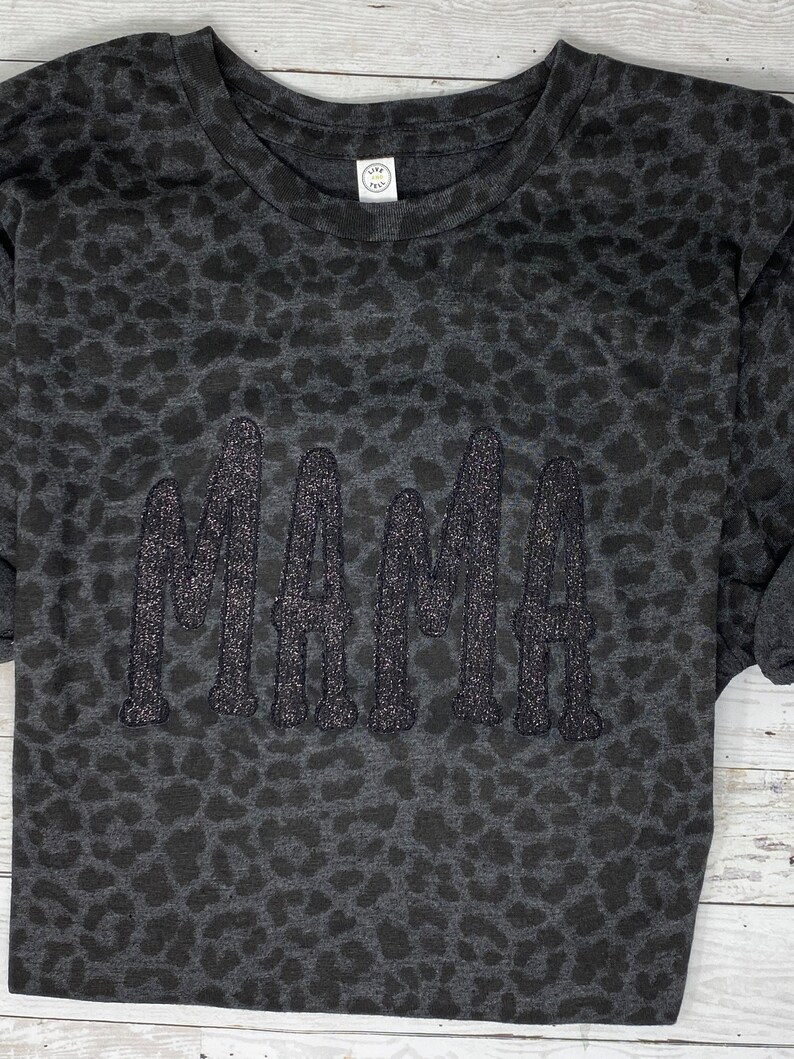 May include: Dark grey sweatshirt featuring a leopard print design. The word "MAMA" is embroidered in glittery, dark grey thread across the front. The sweatshirt has a crew neck and long sleeves.
