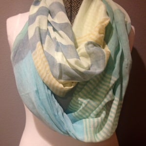 May include: A multi-colored infinity scarf in shades of blue, green, and yellow. The scarf features a striped pattern and is draped over a mannequin. The soft fabric appears lightweight and suitable for warmer weather.