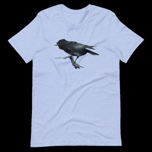 Puede incluir: Una camiseta azul claro con una ilustraci&oacute;n de acuarela negra de un cuervo posado en una rama.