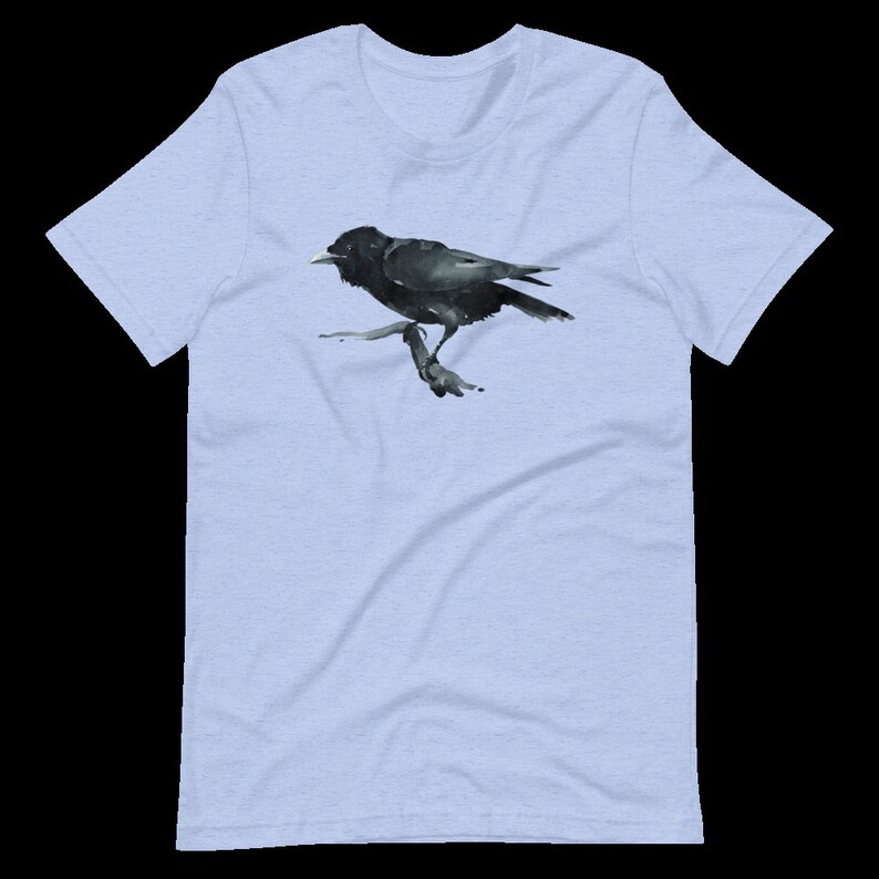Puede incluir: Una camiseta azul claro con una ilustraci&oacute;n de acuarela negra de un cuervo posado en una rama.