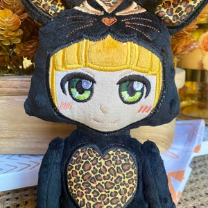 May include: A plush toy with a black and brown leopard print design. The toy is wearing a black hooded costume with a leopard print heart on the front. The hood has cat ears and a cat face embroidered on it. The toy has yellow hair and green eyes.