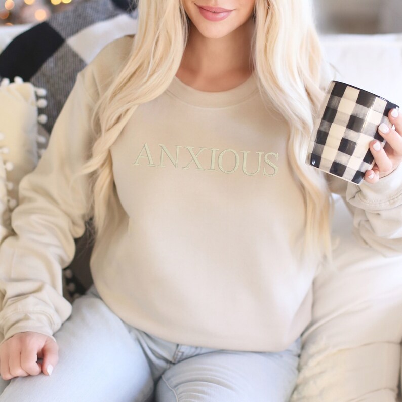 May include: A beige sweatshirt with the word "ANXIOUS" embroidered in green on the front. The person wearing the sweatshirt is holding a black and white checkered mug.