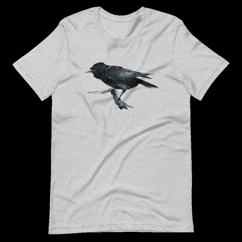 Puede incluir: Una camiseta gris con una ilustraci&oacute;n de cuervo negro impresa en la parte delantera. El cuervo est&aacute; posado en una rama y mira hacia la derecha.