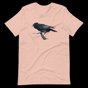 Puede incluir: Una camiseta rosa p&aacute;lido con una ilustraci&oacute;n de cuervo negro. El cuervo est&aacute; posado en una rama y mira hacia la derecha.