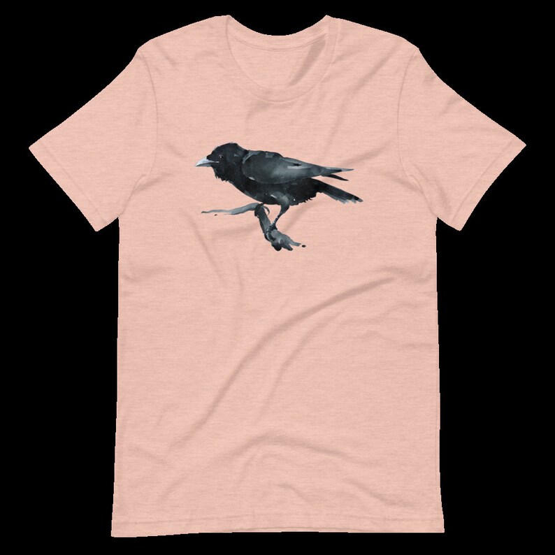 Puede incluir: Una camiseta rosa p&aacute;lido con una ilustraci&oacute;n de cuervo negro. El cuervo est&aacute; posado en una rama y mira hacia la derecha.