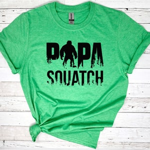 May include: Green t-shirt with a black graphic of a Bigfoot silhouette and the text "Papa Squatch".
