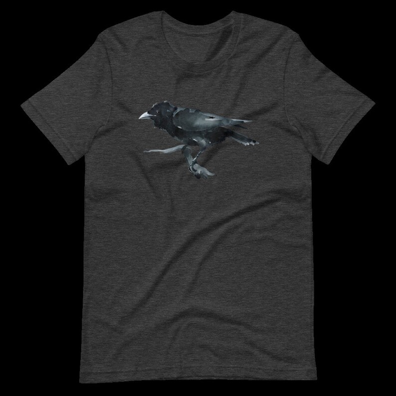 Puede incluir: Una camiseta gris oscuro jaspeada con una ilustraci&oacute;n de cuervo negro. El cuervo est&aacute; posado en una rama y tiene las alas extendidas.