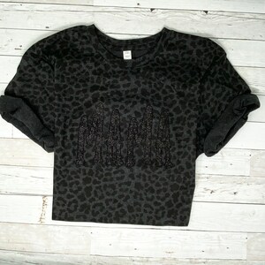 May include: A dark grey t-shirt featuring a leopard print design. The word "MAMA" is embroidered across the front in a darker grey, textured font. The sleeves are cuffed. The shirt is displayed on a white wooden surface.