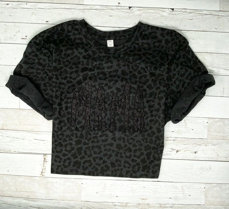 May include: A dark grey t-shirt featuring a leopard print design. The word "MAMA" is embroidered across the front in a darker grey, textured font. The sleeves are cuffed. The shirt is displayed on a white wooden surface.
