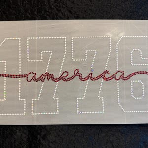 May include: A rectangular patch with the number "1776" outlined in white rhinestones. The word "america" is written in red rhinestones across the center. The background is a light gray fabric.