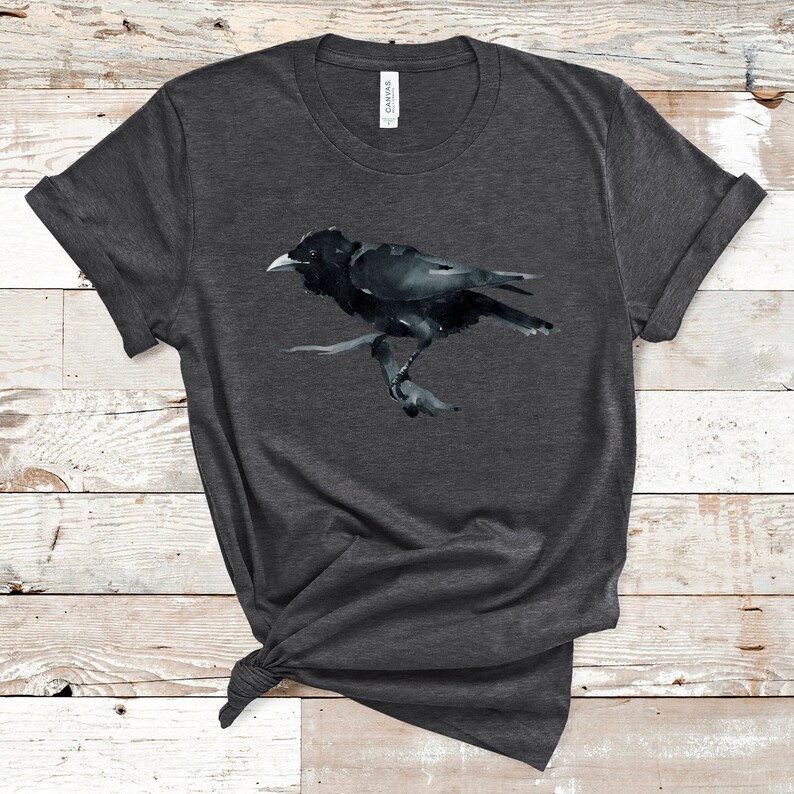 Puede incluir: Una camiseta gris oscuro jaspeada con una ilustraci&oacute;n de acuarela de un cuervo negro.