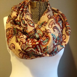 May include: A beige infinity scarf with a paisley pattern in shades of brown, red, teal, and orange. The scarf is draped around a white mannequin, showcasing its design and texture.