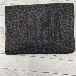 May include: A folded dark grey blanket featuring a leopard print design. The word "MAMA" is embroidered in large, glittery black letters. The blanket is displayed on a light wood surface.