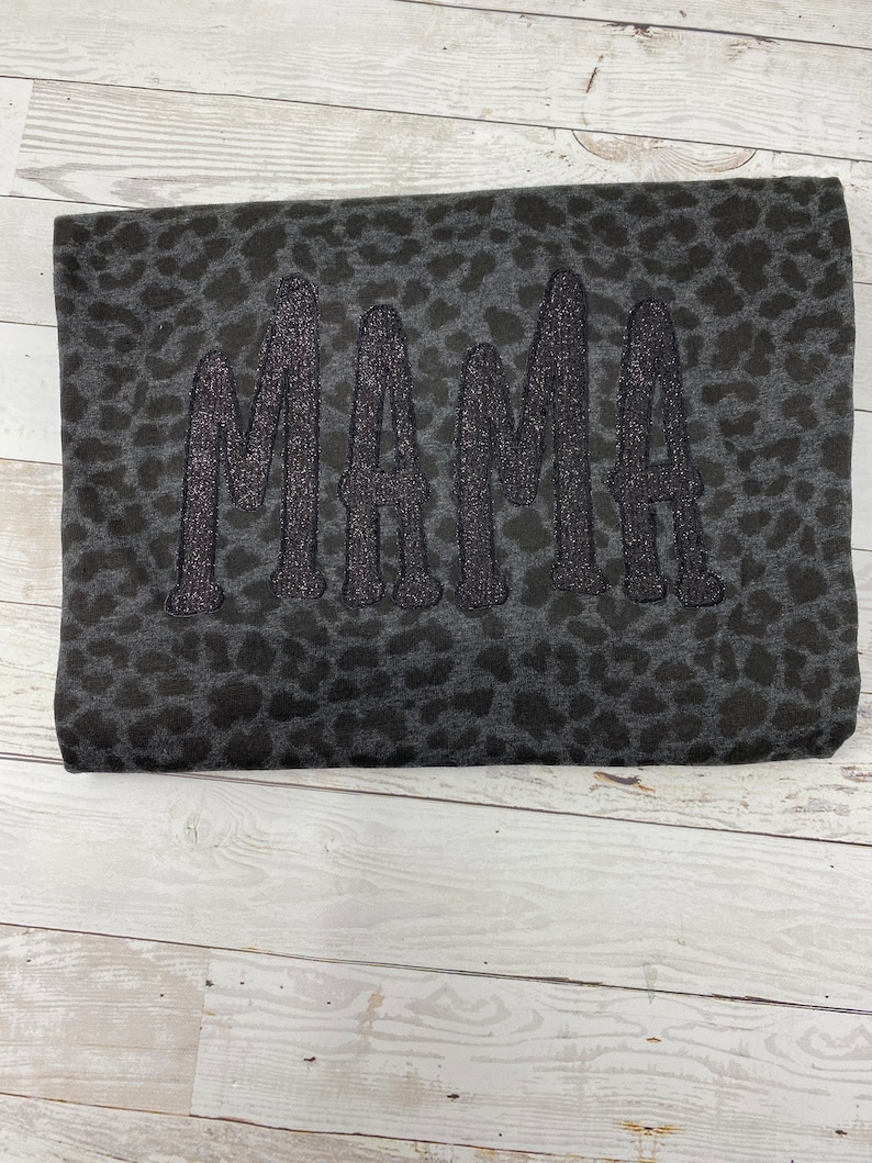 May include: A folded dark grey blanket featuring a leopard print design. The word "MAMA" is embroidered in large, glittery black letters. The blanket is displayed on a light wood surface.