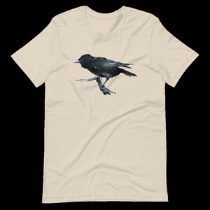 Puede incluir: Una camiseta beige con una ilustraci&oacute;n de cuervo negro. El cuervo est&aacute; posado en una rama y mira hacia la izquierda. El cuervo tiene un pico negro y ojos negros.