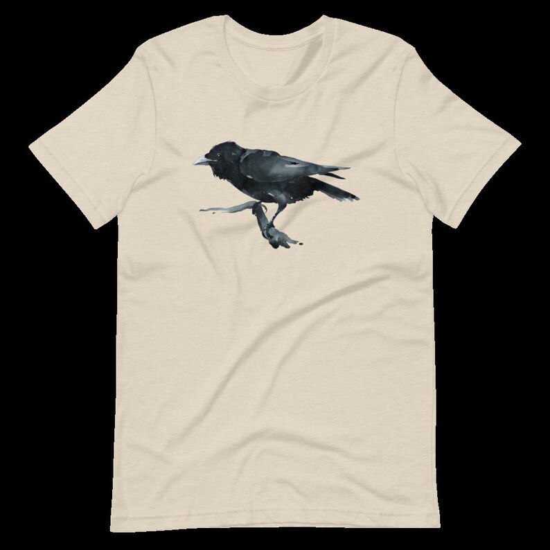 Puede incluir: Una camiseta beige con una ilustraci&oacute;n de cuervo negro. El cuervo est&aacute; posado en una rama y mira hacia la izquierda. El cuervo tiene un pico negro y ojos negros.