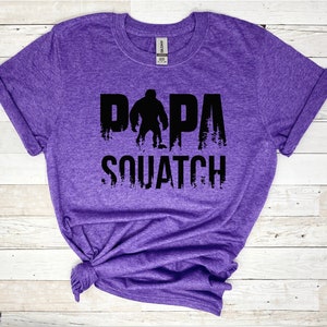 May include: Purple t-shirt with a black graphic of a Bigfoot with the text "Papa Squatch".