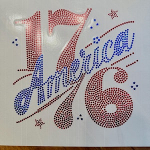May include: A rhinestone transfer design on a white backing featuring the number 17 in red, the word America in blue script, and the number 6 in red. The design is accented with small blue and red stars and dots, creating a patriotic theme.