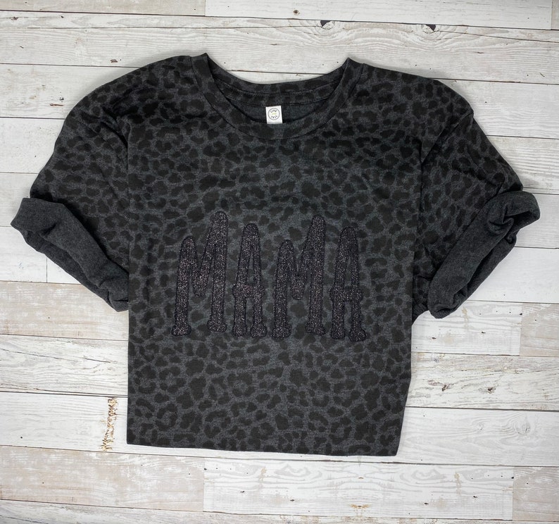 May include: A dark grey t-shirt featuring a leopard print design. The word "MAMA" is embroidered in glittery dark grey thread across the front. The sleeves are rolled up.
