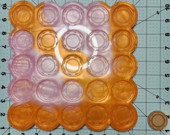 Puede incluir: Un molde de resina rosa y dorado con 25 impresiones circulares. El molde está diseñado para crear piezas circulares de resina con un borde elevado.
