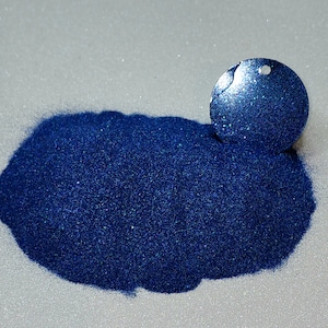 Puede incluir: Un montón de purpurina azul oscuro con brillo iridiscente. Se muestra una pequeña muestra de la purpurina en un recipiente redondo.