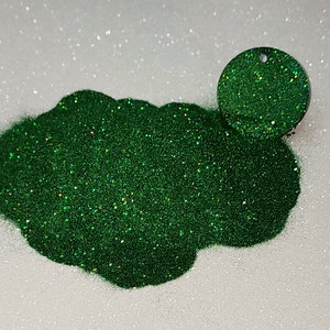 Puede incluir: Primer plano de purpurina verde con brillo iridiscente. La purpurina está amontonada en un montículo suelto sobre una superficie blanca.