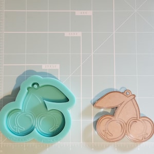 May include: A light blue silicone mold of a cherry with a stem and leaf. The mold is on a green gridded surface. A brown wooden cutout of a cherry with a stem and leaf is on the right side of the image.