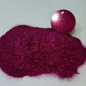 Puede incluir: Un montón de purpurina magenta brillante junto a un dije redondo a juego con un agujero. La purpurina está finamente molida y refleja la luz, creando un efecto deslumbrante. El fondo es una superficie blanca texturizada.