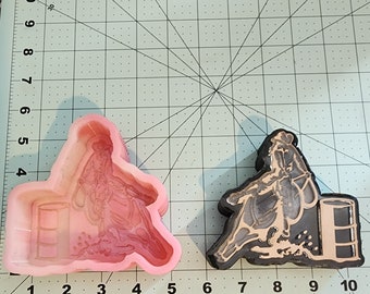 Puede incluir: Un molde de silicona rosa de un jinete de barriles en un caballo, con un barril en el fondo. El molde está en una estera de corte verde con marcas en pulgadas.