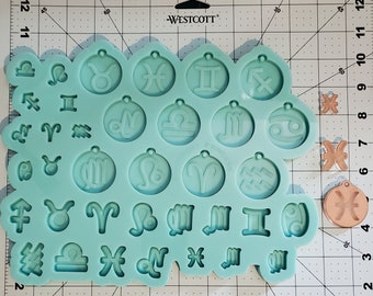May include: A light blue silicone mold with 24 different zodiac signs. The mold is on a white gridded surface.