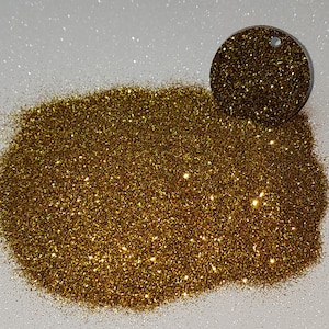 Puede incluir: Un montón de brillantina dorada fina con brillo iridiscente. La brillantina está suelta y extendida sobre una superficie blanca.