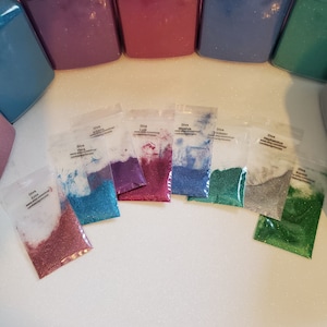 Puede incluir: Una variedad de purpurina en varios colores, incluyendo rosa, azul, morado y verde, exhibida en pequeñas bolsas de plástico transparente. Las bolsas están etiquetadas con la palabra "Diva" y otros nombres. Contenedores más grandes de purpurina en colores a juego son visibles.