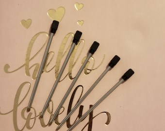 May include: Five silver pen refills with black tips are arranged diagonally across a pink surface. The surface has gold foil hearts and the words "hello lovely" in cursive script.