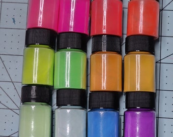 Könnte beinhalten: Zwölf kleine Glasflaschen mit schwarzen Deckeln, gefüllt mit verschiedenen Farben von Neon-Pigmentpulver. Die Flaschen sind in drei Reihen zu vier angeordnet, wobei die Farben von Pink nach Lila übergehen.